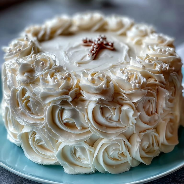 Classic vanilla sponge cake decorated with silky buttercream, delicate white roses, and a silver cross topper for a timeless First Communion celebration.