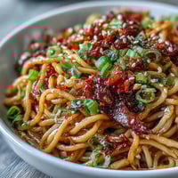 A steaming bowl of chili crisp noodles, glistening with chili oil and topped with fresh scallions and crunchy peanuts.